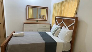 Una cama bien hecha con colcha gris y blanca, cabecera de madera y mesita de noche con un espejo y una pequeña planta.