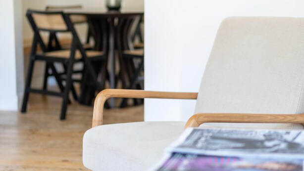 Una sala de estar moderna con un sofá de color claro y apoyabrazos de madera. Al fondo, se observa un comedor con sillas de madera oscura y una mesa.