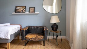 Un dormitorio con cama, un sillón de cuero negro, una mesita de noche con lámpara, un espejo y una estantería con dos cuadros enmarcados.