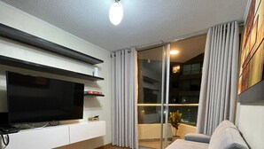 Un soggiorno moderno con televisione a schermo piatto, pavimento in legno, un divano grigio e una porta vetrata scorrevole che dà su un balcone.