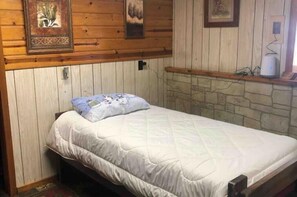 A single bed with a white quilted mattress and a blue pillow with a cartoon character. The room has wooden panel walls and a stone wall at the bottom. - Country Crossroads (Armstrong Creek)