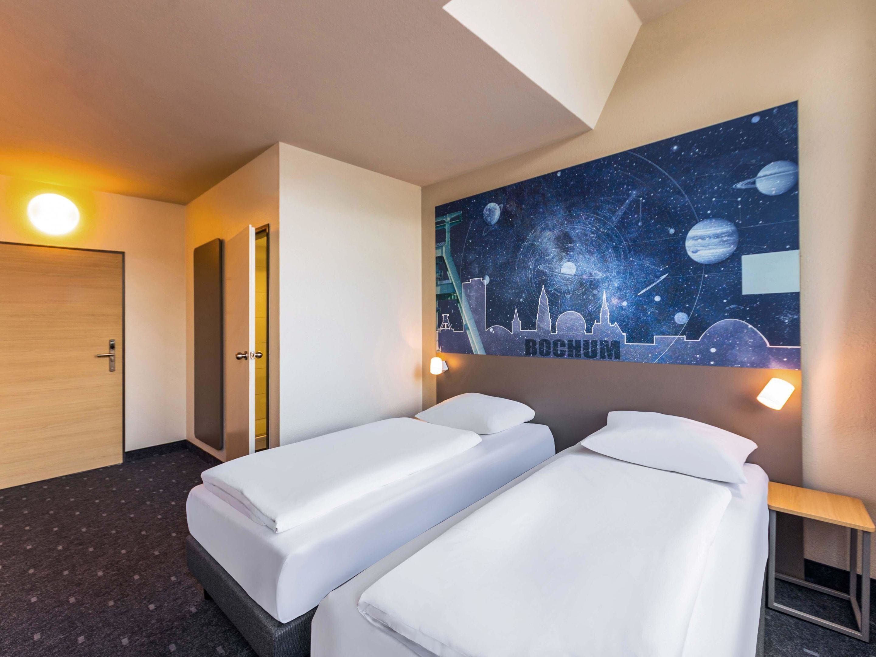 Une chambre d’hôtel avec deux lits simples, une fresque représentant un ciel étoilé sur le mur et une porte en bois.