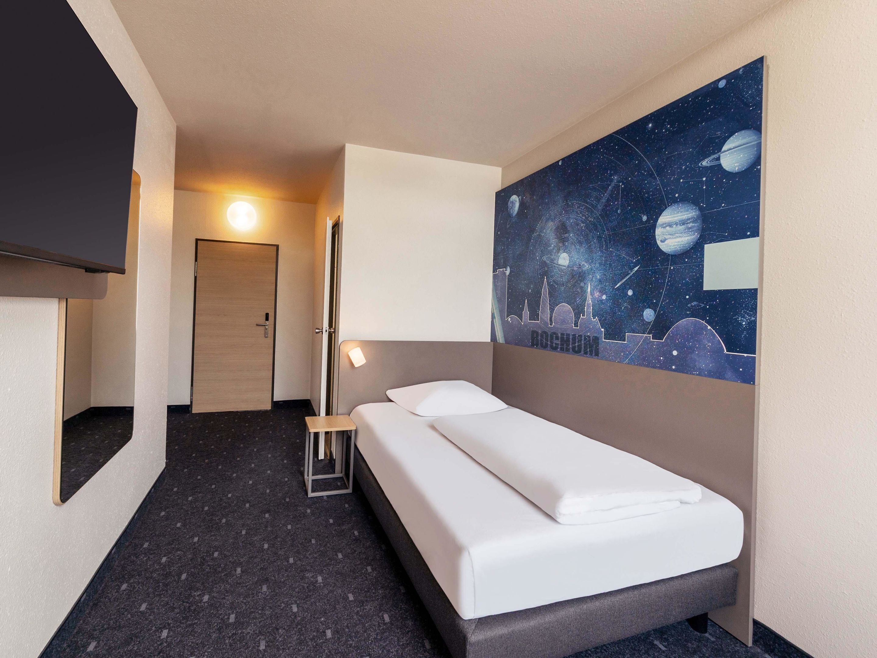 Ein Hotelzimmer mit Bett, Nachttisch, einem an der Wand befestigten Fernseher und einem Bild mit Weltraumdarstellung.