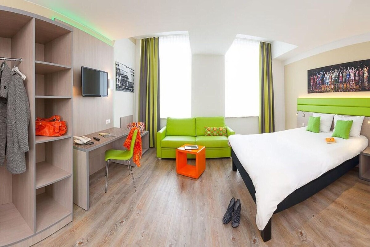 A modern hotel room with a bed, a desk, a green sofa, and a wardrobe.