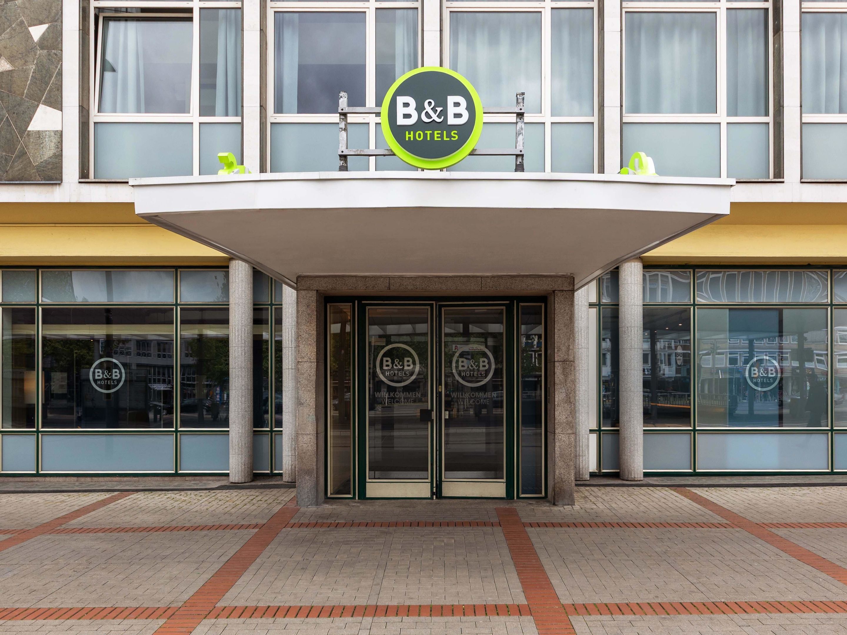 Entrada de um hotel B&B com design moderno, com grandes portas de vidro e o logo B&B bem visível acima da entrada.