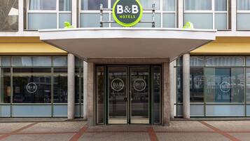 Entrada de um hotel B&B com design moderno, com grandes portas de vidro e o logo B&B bem visível acima da entrada.