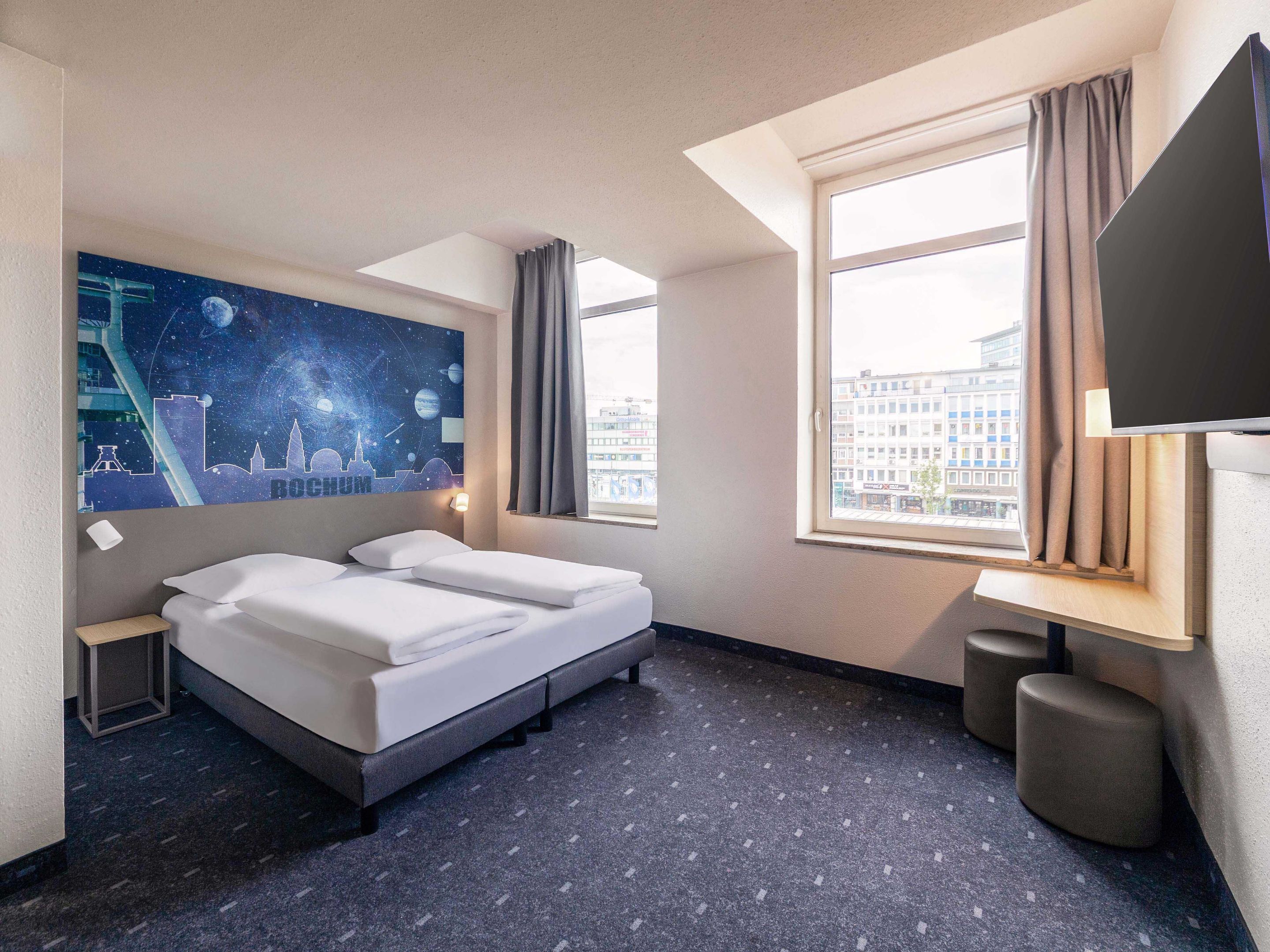 Una habitación de hotel con cama, un cuadro de arte urbano en la pared, una ventana con cortinas y un televisor de pantalla plana.