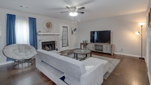 Een woonkamer met een witte bank, een ronde salontafel, een open haard, een plafondventilator en een flatscreen televisie.