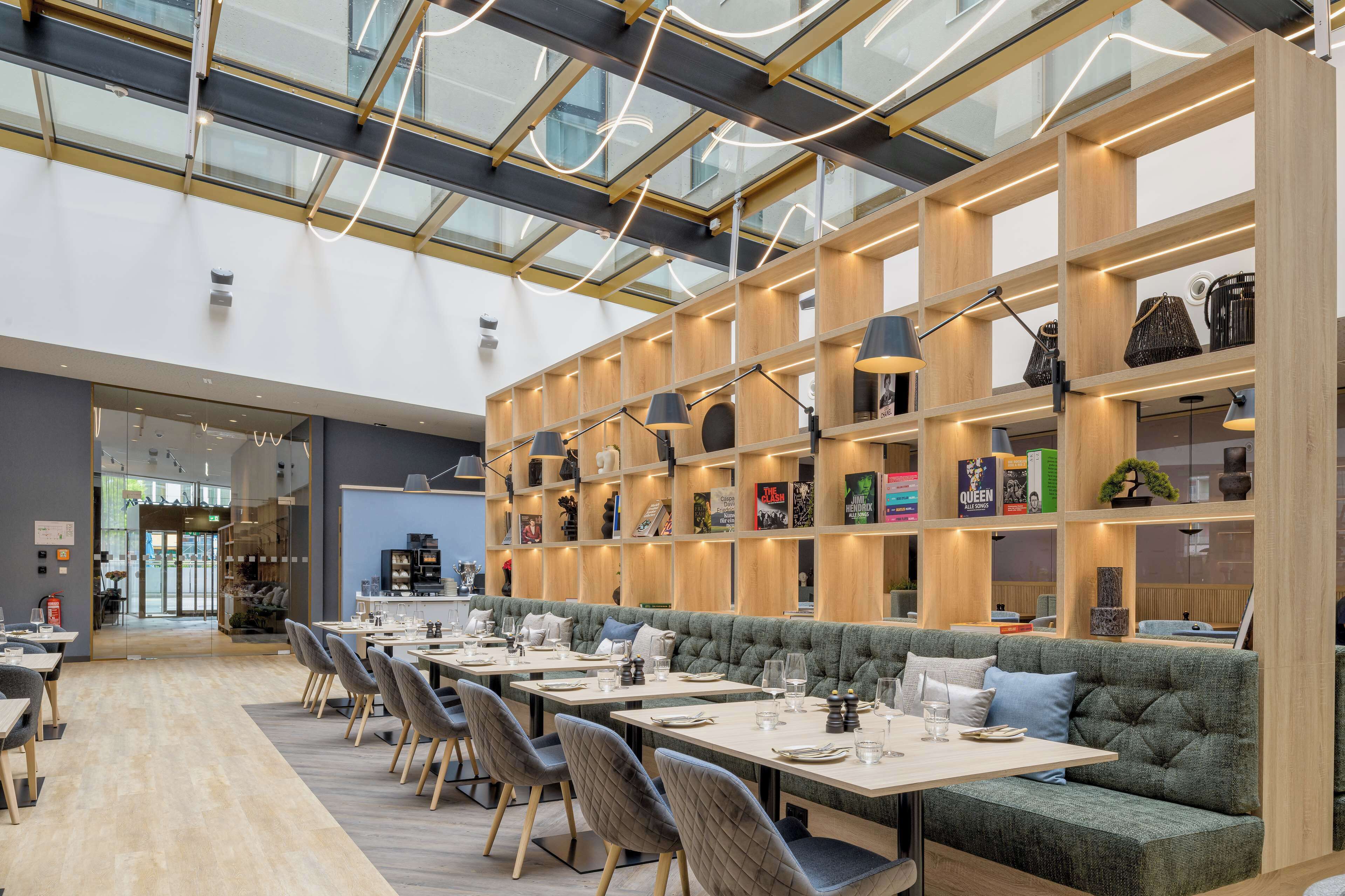 Un ristorante moderno con soffitto alto, pannelli di vetro ed scaffali in legno con libri e oggetti decorativi.