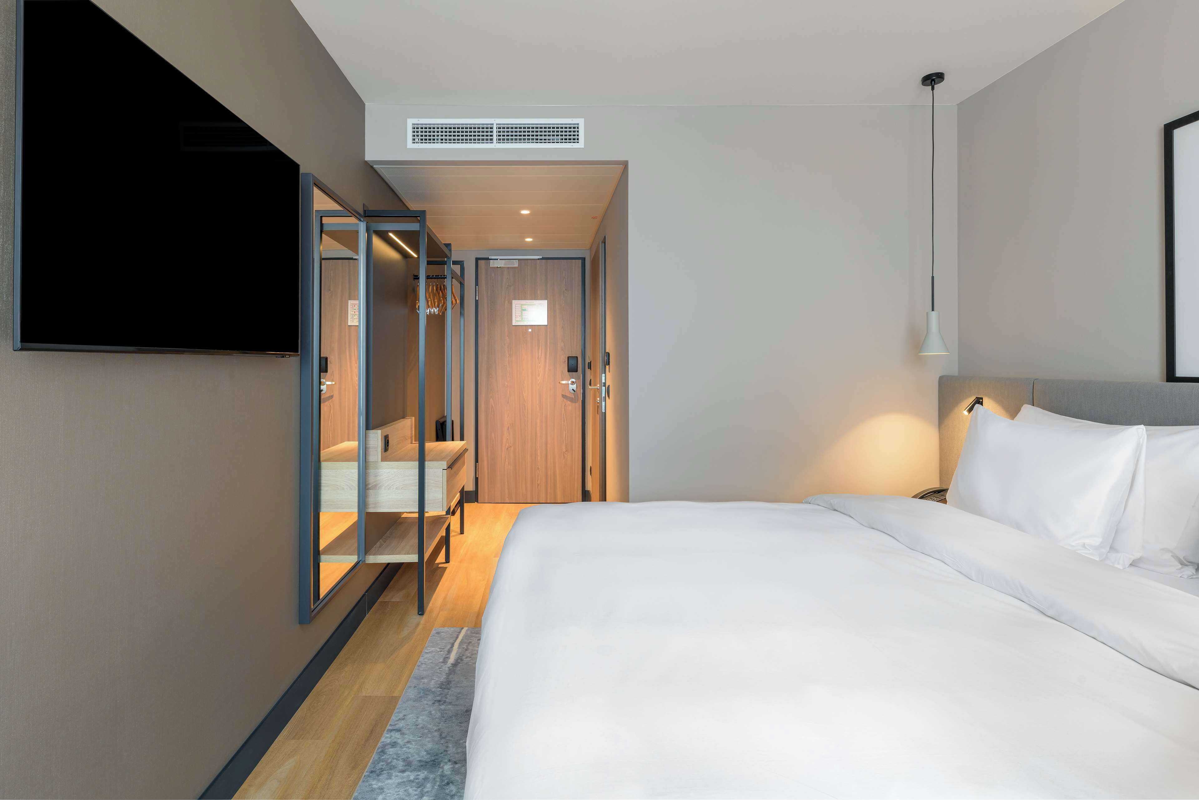 Una habitación de hotel moderna con una cama grande, un televisor de pantalla plana montado en la pared, un vestidor y una mesita de noche con una lámpara.