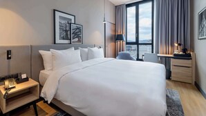 A modern hotel room with a large bed, a desk, and a chair. There is a window with curtains, a wall-mounted phone, and framed pictures.