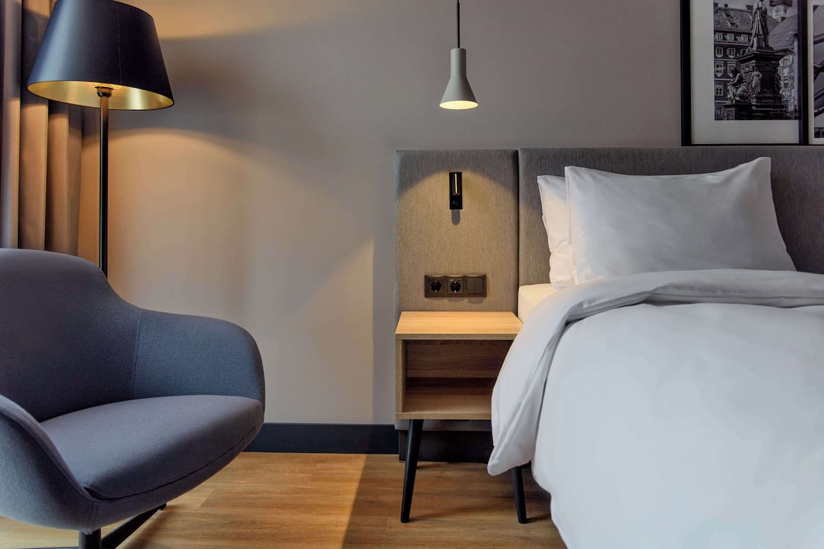 A hotel room with a bed, a chair, a bedside table, a lamp, and a wall-mounted light fixture.