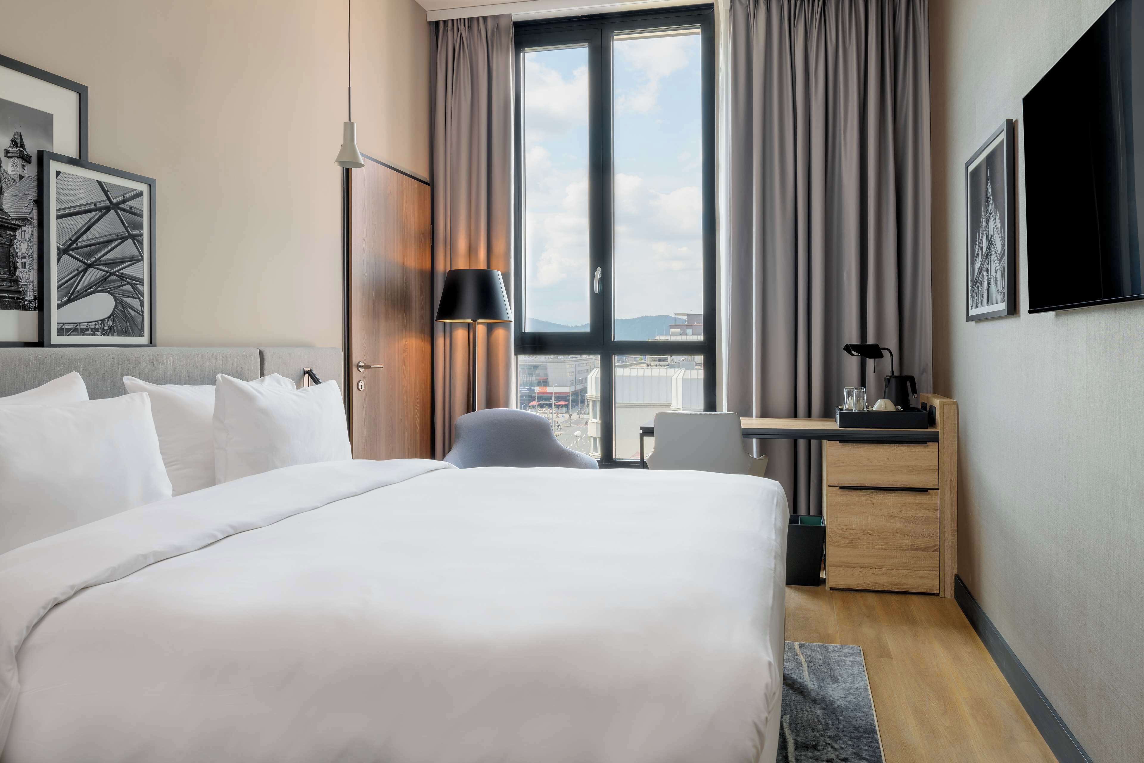 A hotel room with a large bed, a desk, a chair, and a TV on the wall.