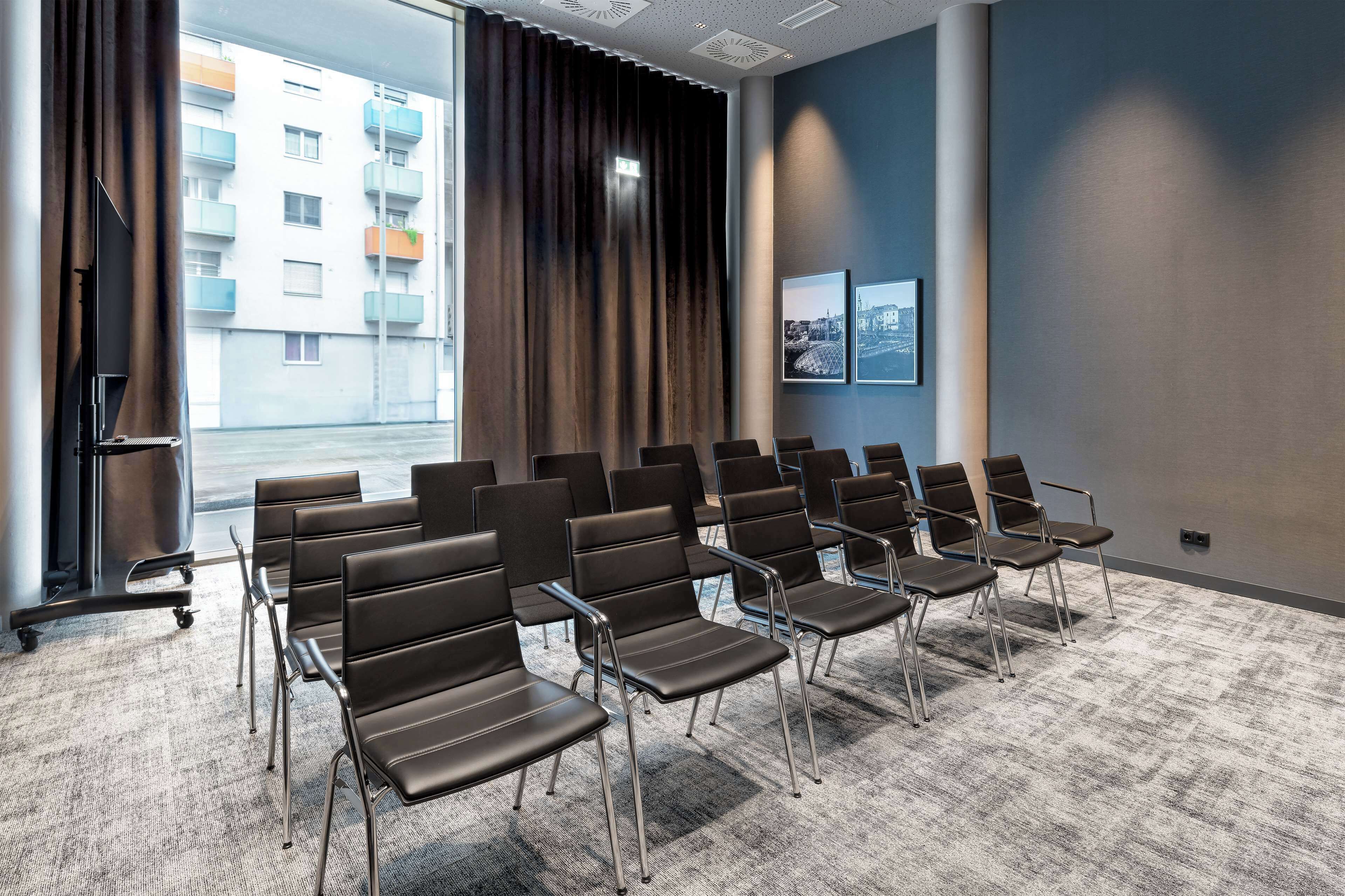 Een congreszaal met rijen zwarte stoelen, een groot raam met gordijnen en een vloerbedekking.