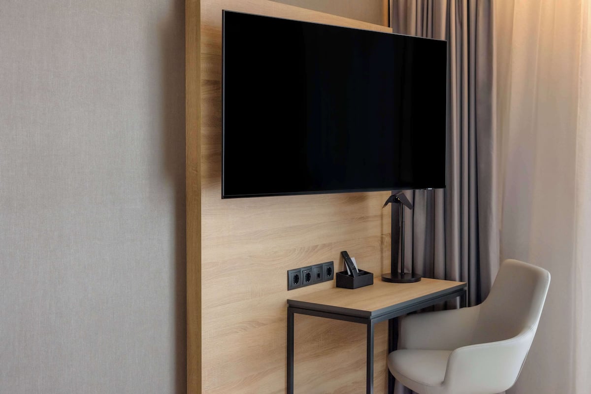 A modern hotel room with a flat-screen TV mounted on a wooden wall, a small table with a lamp, and a white chair.