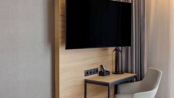 A modern hotel room with a flat-screen TV mounted on a wooden wall, a small table with a lamp, and a white chair.