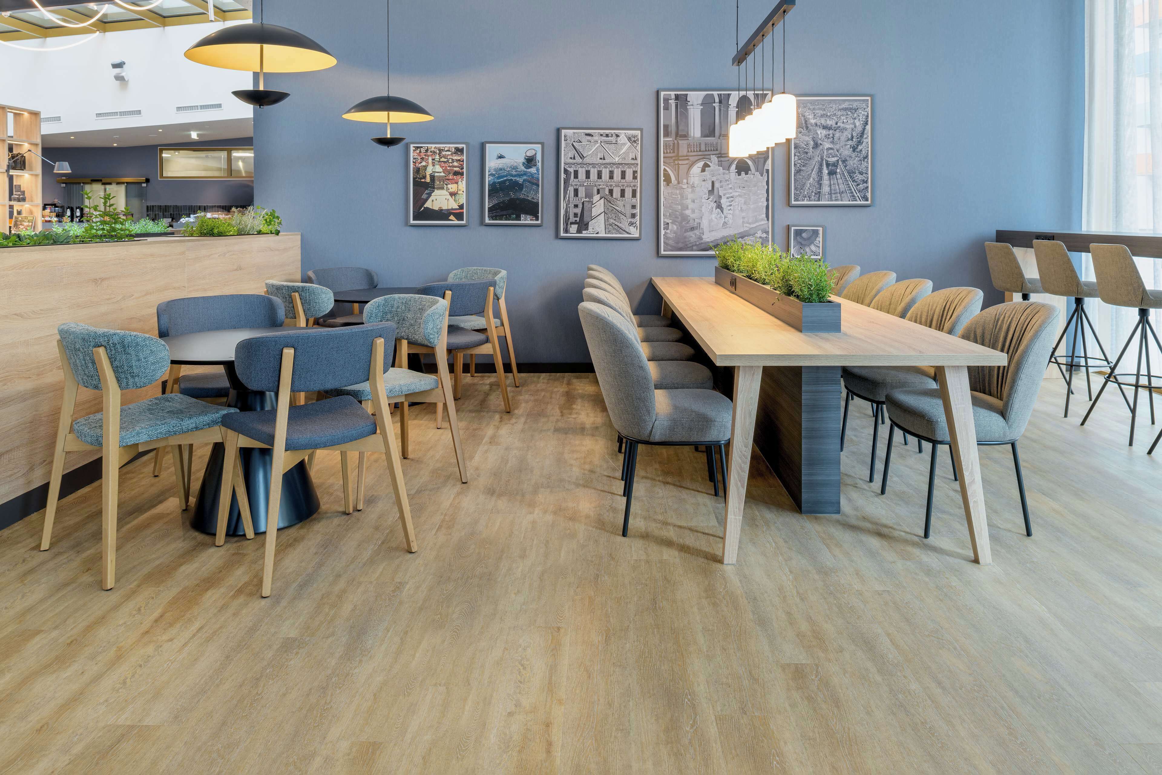 Un espace repas moderne avec une longue table et de nombreuses chaises, une cuisine en arrière-plan et des tableaux représentant des paysages urbains encadrés au mur.