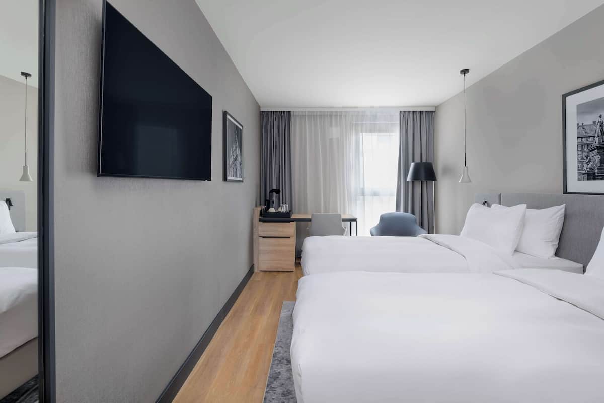 A hotel room with two beds, a desk, a chair, a TV, and a window with curtains.