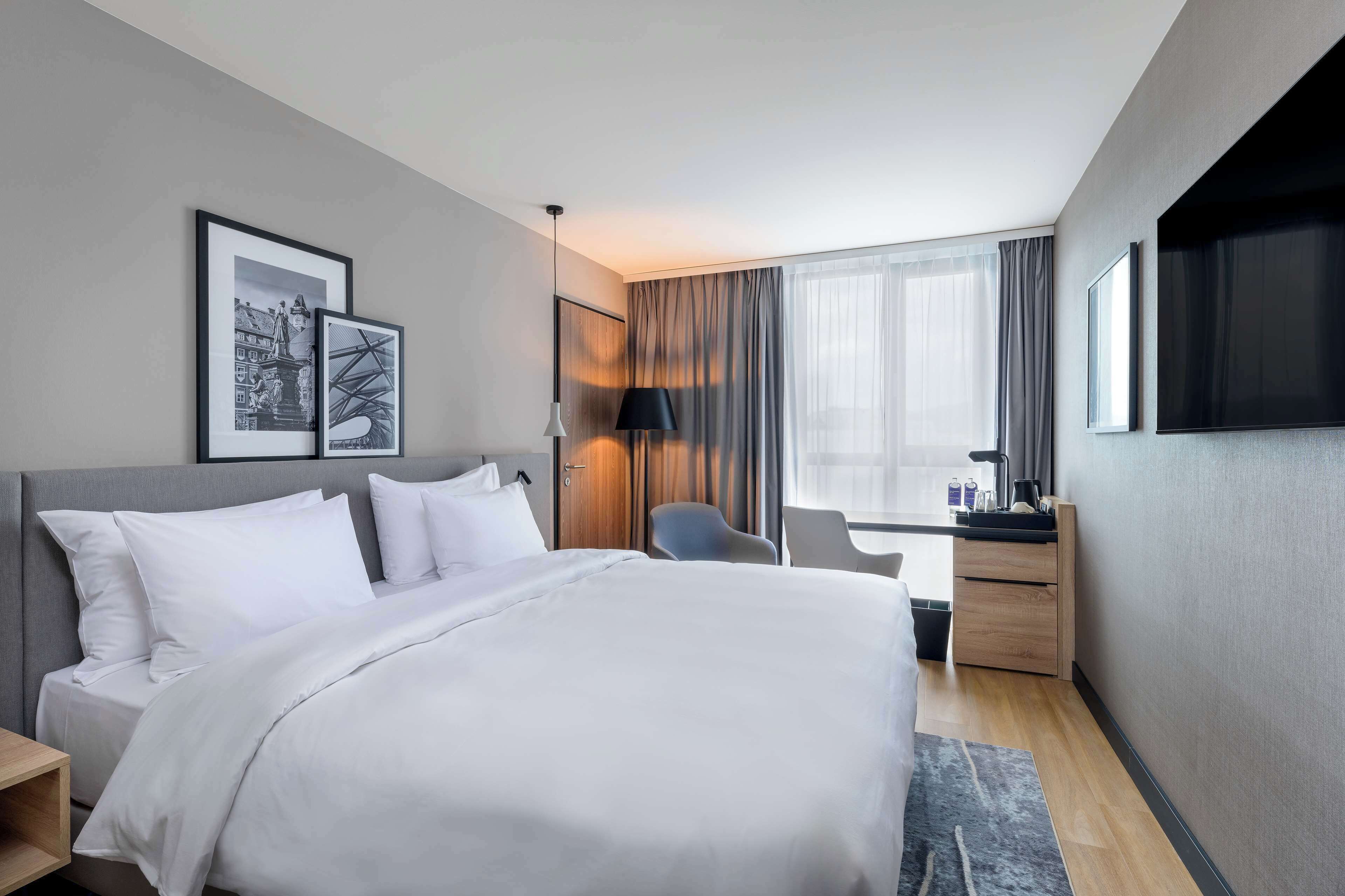 A modern hotel room with a large bed, a desk, a chair, and a TV.