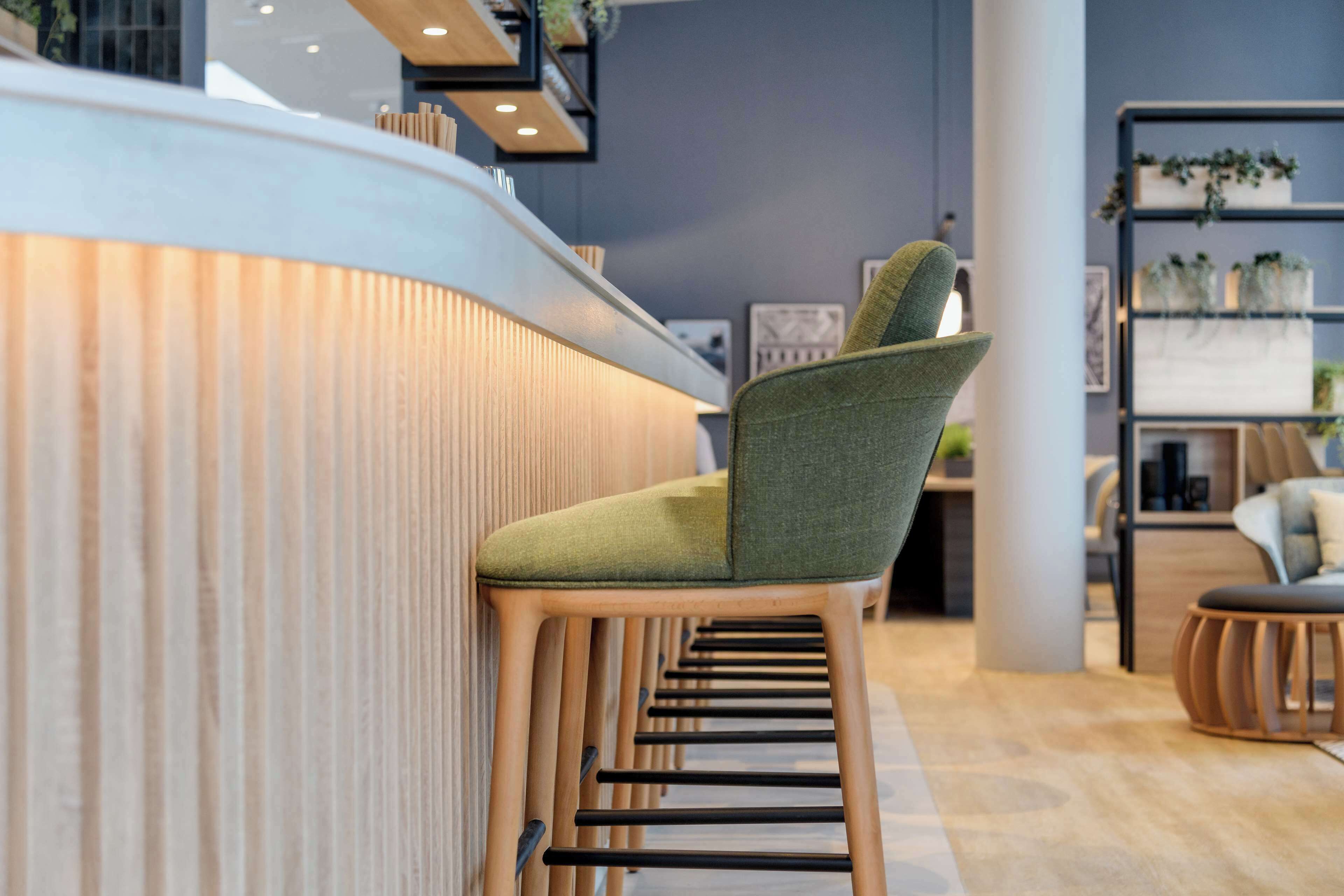 A modern bar area with a green upholstered bar stool, wooden counter, and shelves with plants.