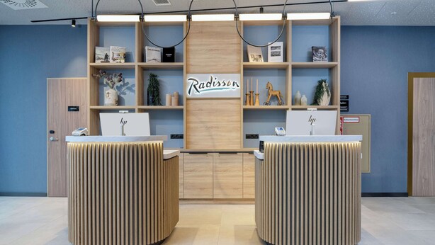A reception area with two wooden reception desks, a central wooden panel with the name 'Radisson', and shelves with various items.