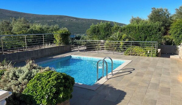 Una piscina rectangular con escaleras metálicas, rodeada por un solárium de baldosas y barandillas de metal, con montañas al fondo.