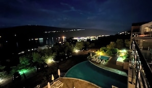 Vista nocturna de un resort con piscina, sillones de descanso y un paisaje urbano distante.