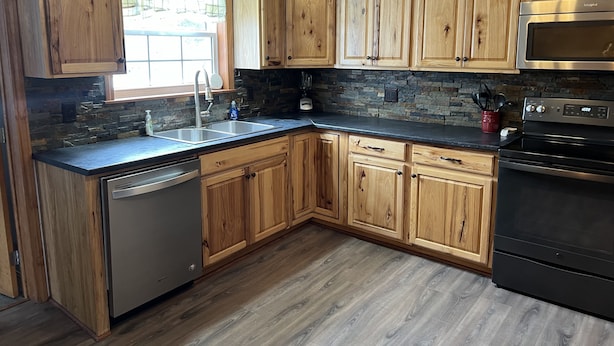 A kitchen with wooden cabinets, a stone backsplash, a stainless steel dishwasher, a black oven, and a window with a view of greenery. - Newly renovated, only 2 miles from downtown Fort Kent.  Right across from trails (Fort Kent)