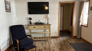 A room with a dark blue armchair, a wooden console table with a lamp, a flat-screen TV, and a door leading to another room. - Newly renovated, only 2 miles from downtown Fort Kent.  Right across from trails (Fort Kent)