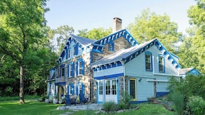A two-story house with a stone facade and blue trim, surrounded by a well-maintained garden and trees. - Spacious 4BD Country Gem - Fire Pit & Chill Vibes (Highland)