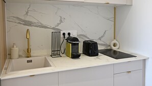 A compact kitchen with white cabinets, a marble backsplash, a gold faucet, a kettle, a coffee maker, and a small sink.