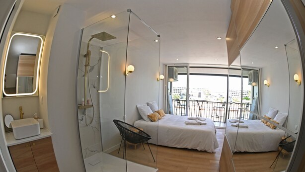 A modern hotel room with a glass shower enclosure, a large mirror, a bed with white linens, and a view of the city through large windows.