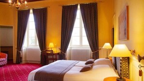A hotel room with a large bed, two windows with curtains, a chandelier, a table with a lamp, and a red carpet. - Château le Haget (Montesquiou)