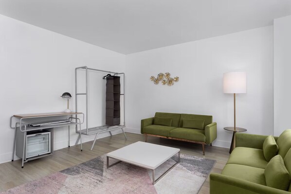 A modern living room with a green sofa, a white coffee table, a metal desk with a lamp, and a wall-mounted shelf.