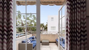 A balcony with wicker furniture and cushions, a view of palm trees, and a building in the background. - Roami at Hotel Astor (Miami Beach)