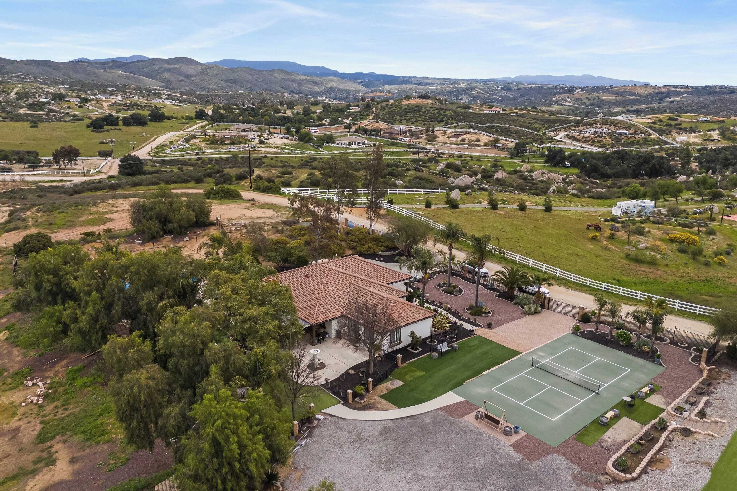 An aerial view of a residential area with a tennis court, a house with a patio, and surrounding greenery.