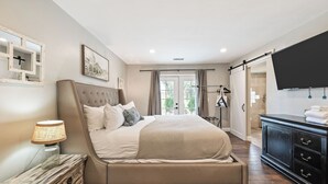 A bedroom with a bed, a bedside lamp, a dresser, a TV, and a door leading to a balcony. - Breathtaking Meadows by Avantstay Temecula Group Retreat Pool, Pickleball, Firepit, Hot Tub (Temecula)