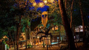 Une cabane perchée dans un arbre, dotée d’un toit de chaume, entourée d’une végétation luxuriante et illuminée par des guirlandes lumineuses.