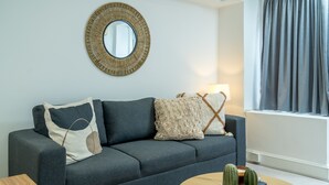 Een moderne woonkamer met een grijze bank, een ronde salontafel met cactussen, een geweven spiegel en een lamp.