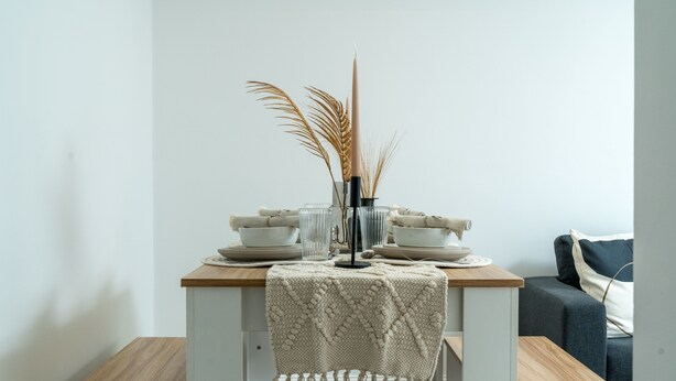Een kleine eettafel met een witte tafelkleed, twee witte borden, een glas en een vaas met gedroogde planten.