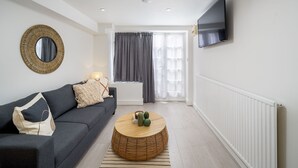 Een moderne woonkamer met een donkergrijze bank, een ronde houten salontafel met decoratieve objecten, een grote spiegel en een aan de muur bevestigde televisie.