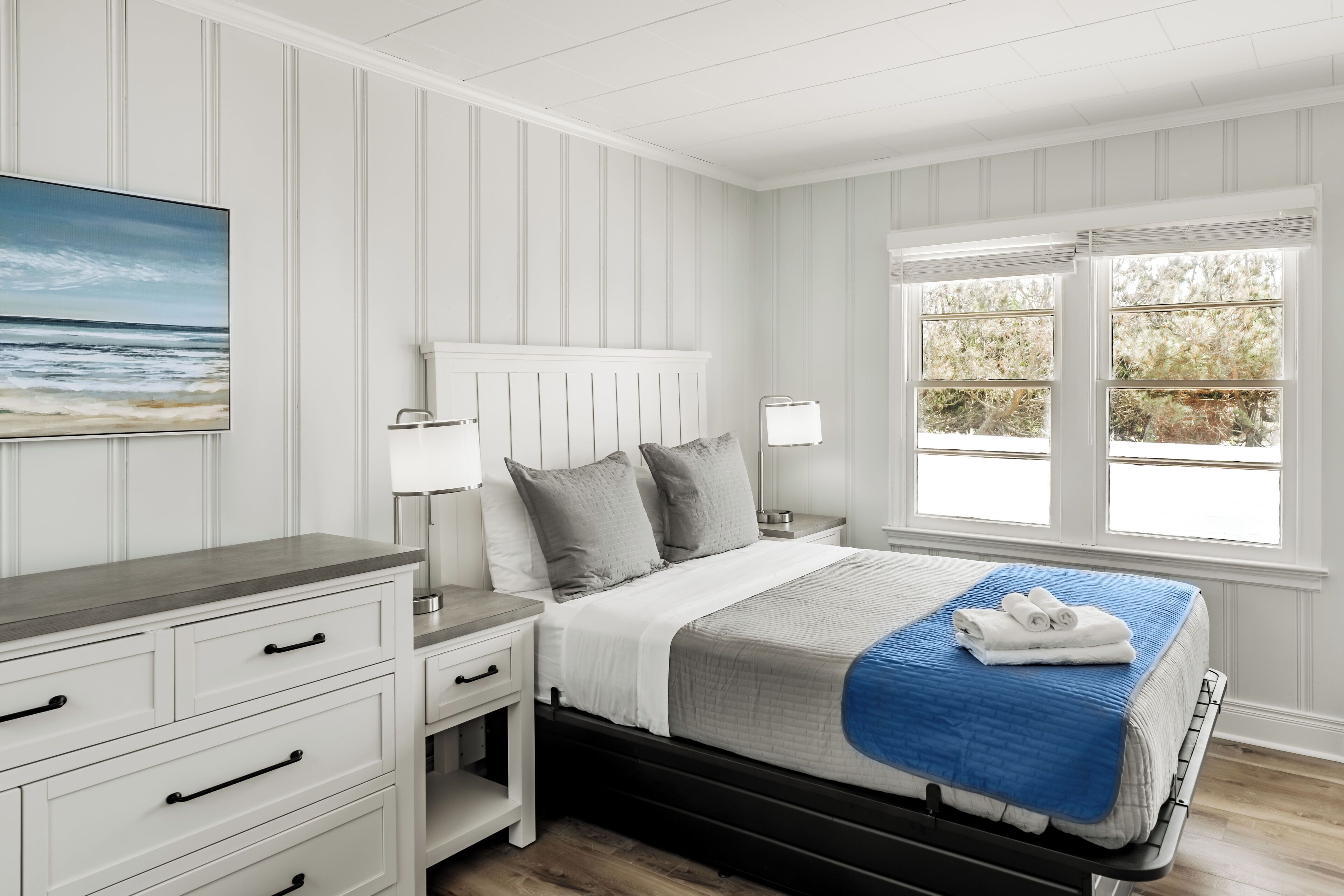 A bedroom with a bed, nightstands, a dresser, and a painting of a beach scene.