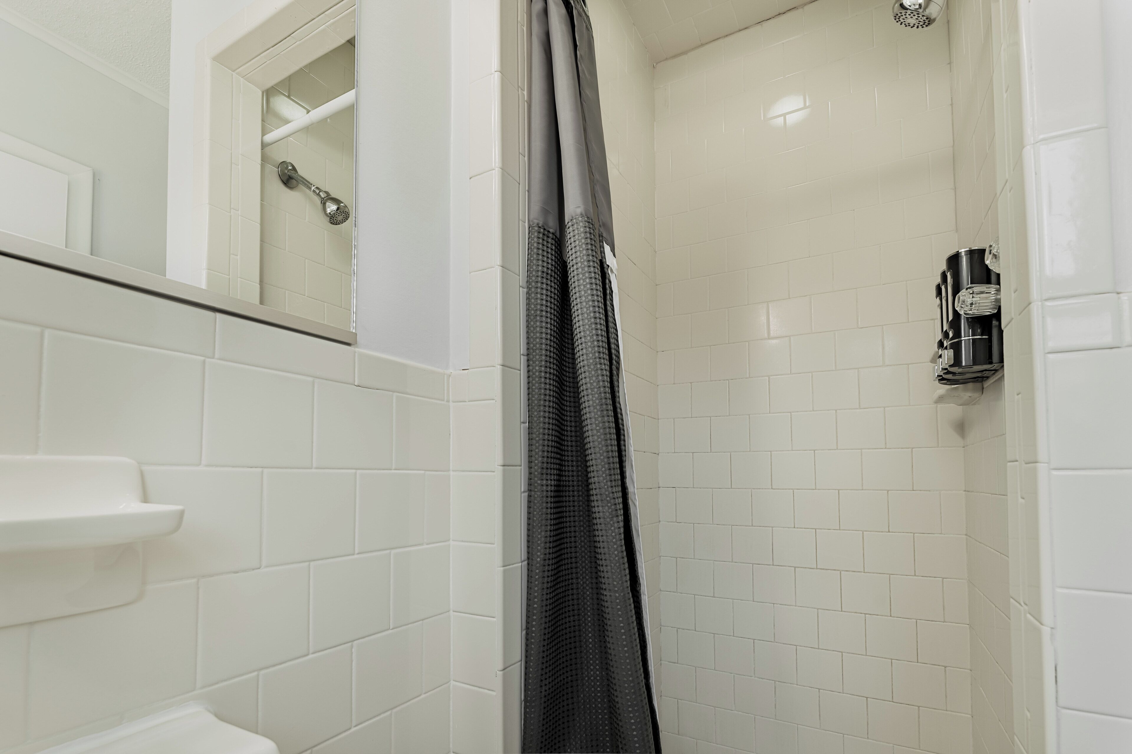 Un baño con paredes de azulejos blancos, una ducha con cortina gris y un lavabo blanco con jabonera.