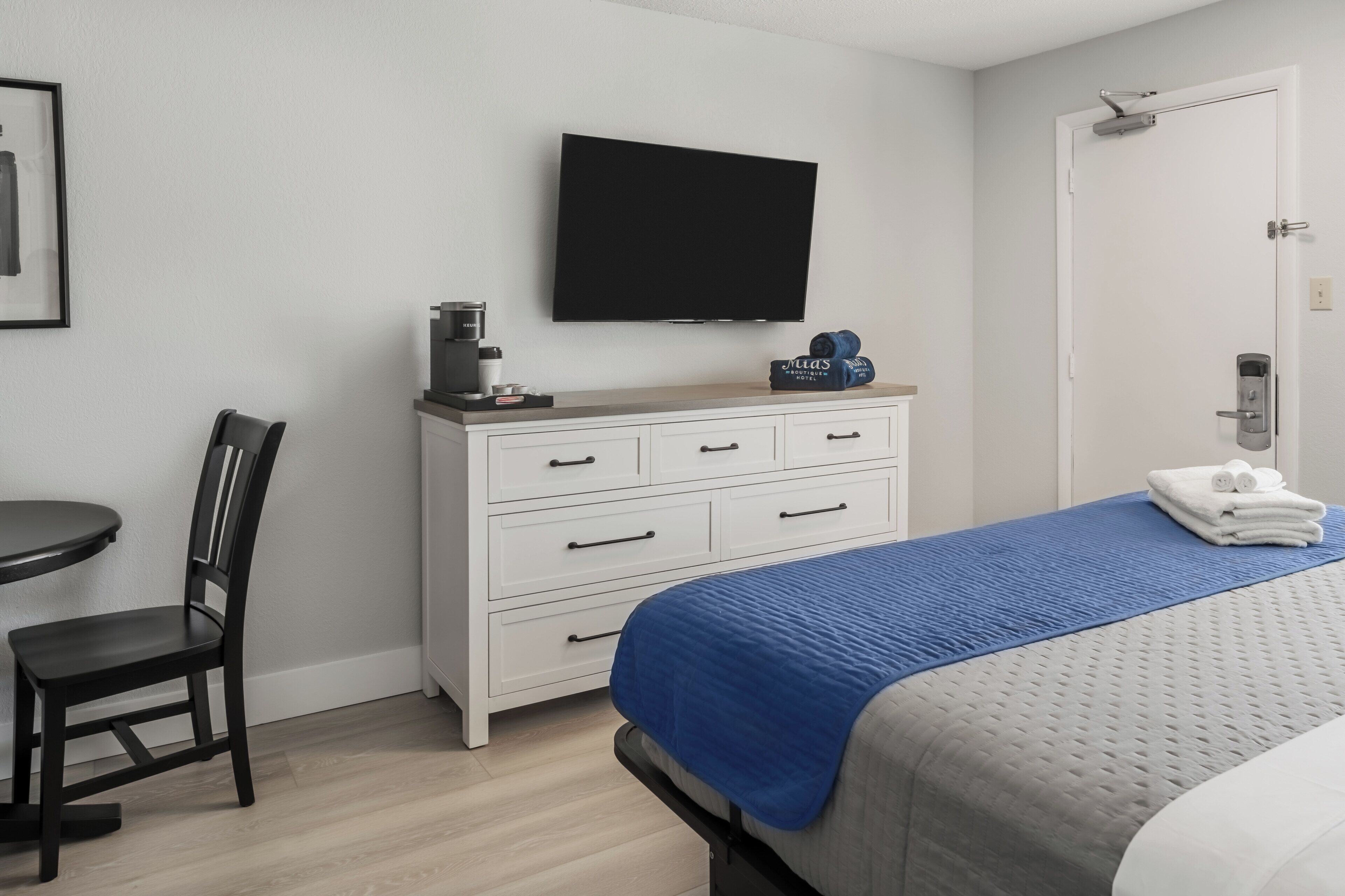 Une chambre d’hôtel comprenant un lit, un bureau avec une chaise, une commode, un téléviseur fixé au mur et une porte avec une serrure.