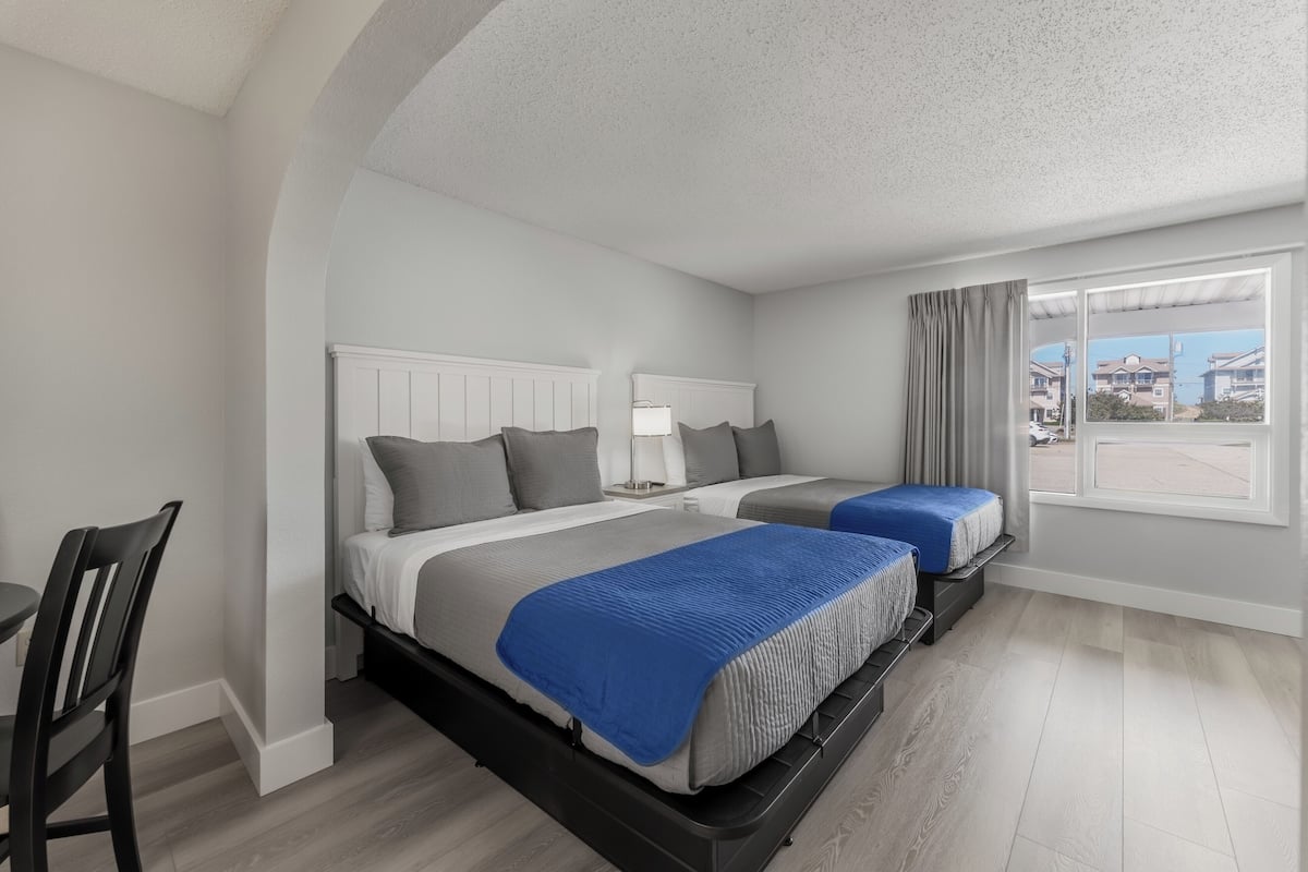 A hotel room with two beds, a desk, and a window with a view of buildings.