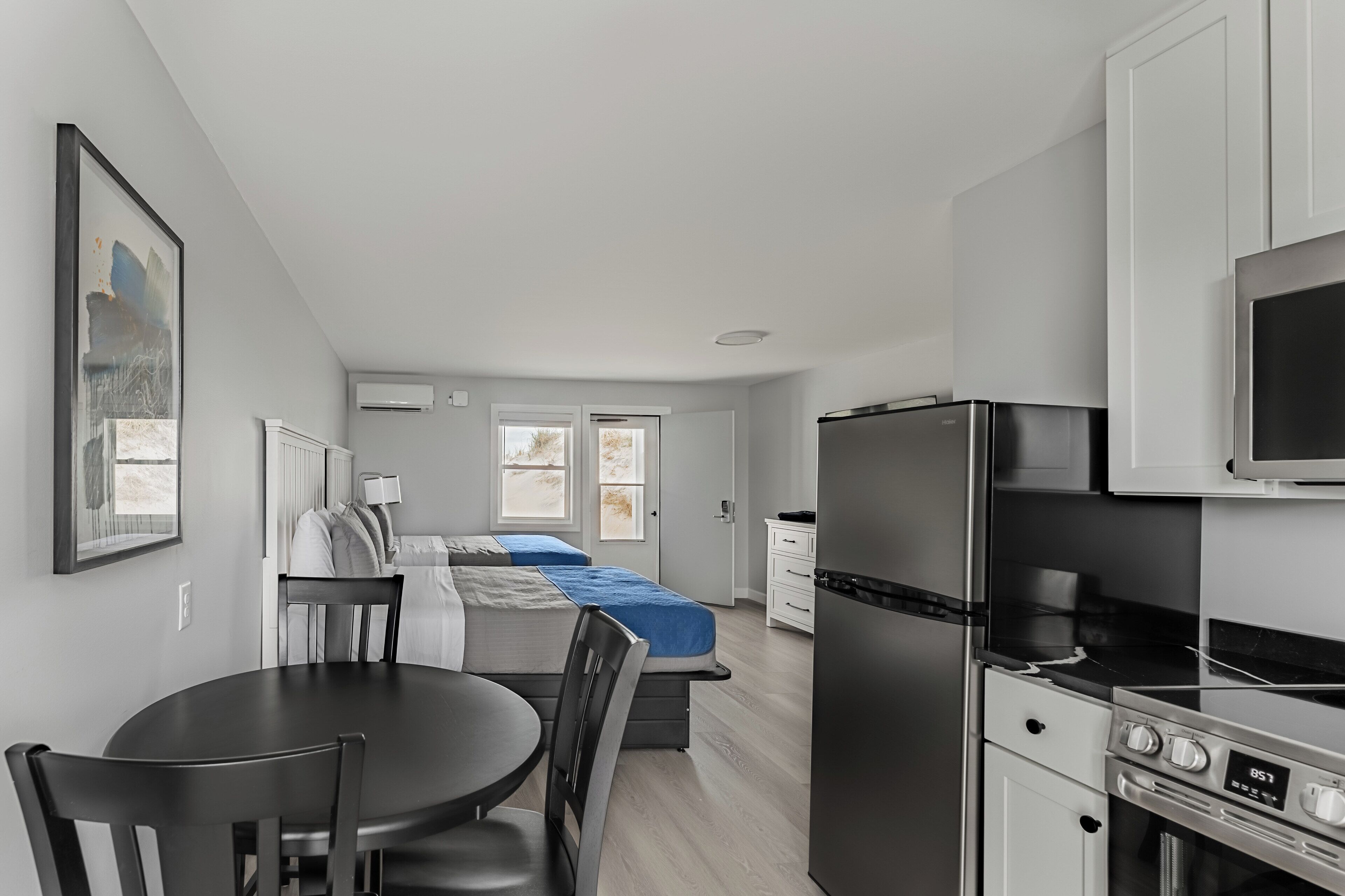 Cocina compacta con comedor, cama con cubrecama azul y un ventanal grande.