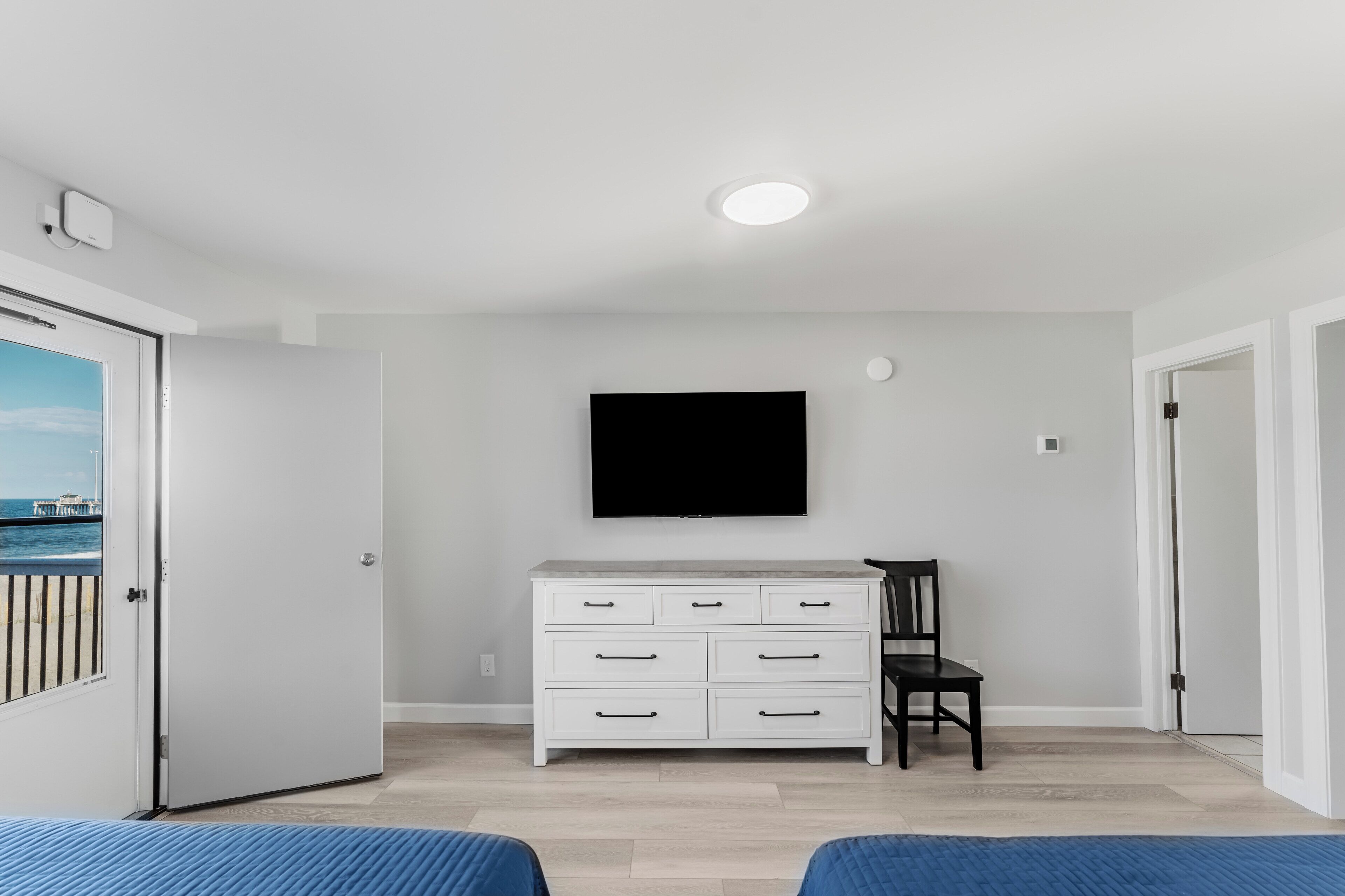 A room with a white dresser, a flat-screen TV mounted on the wall, a chair, and a bed with a blue bedspread.