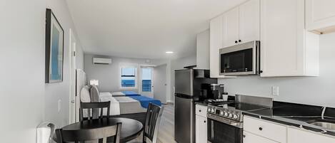 A modern kitchen with white cabinets, a stainless steel refrigerator, a microwave, and a stove. There is a dining area with a table and chairs.