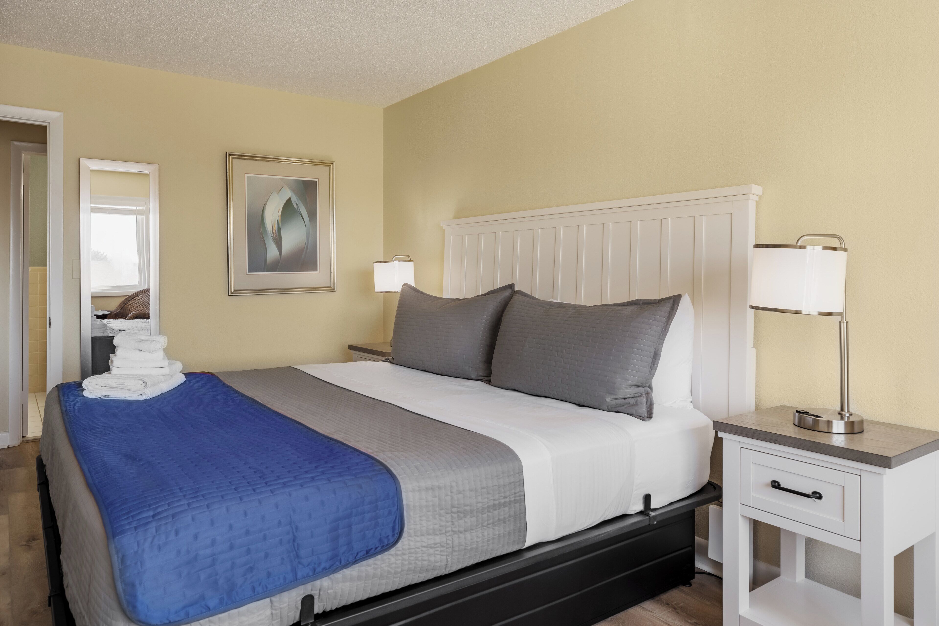 A neatly made bed with a blue and white bedspread, a white nightstand with a lamp, and a framed picture on the wall.