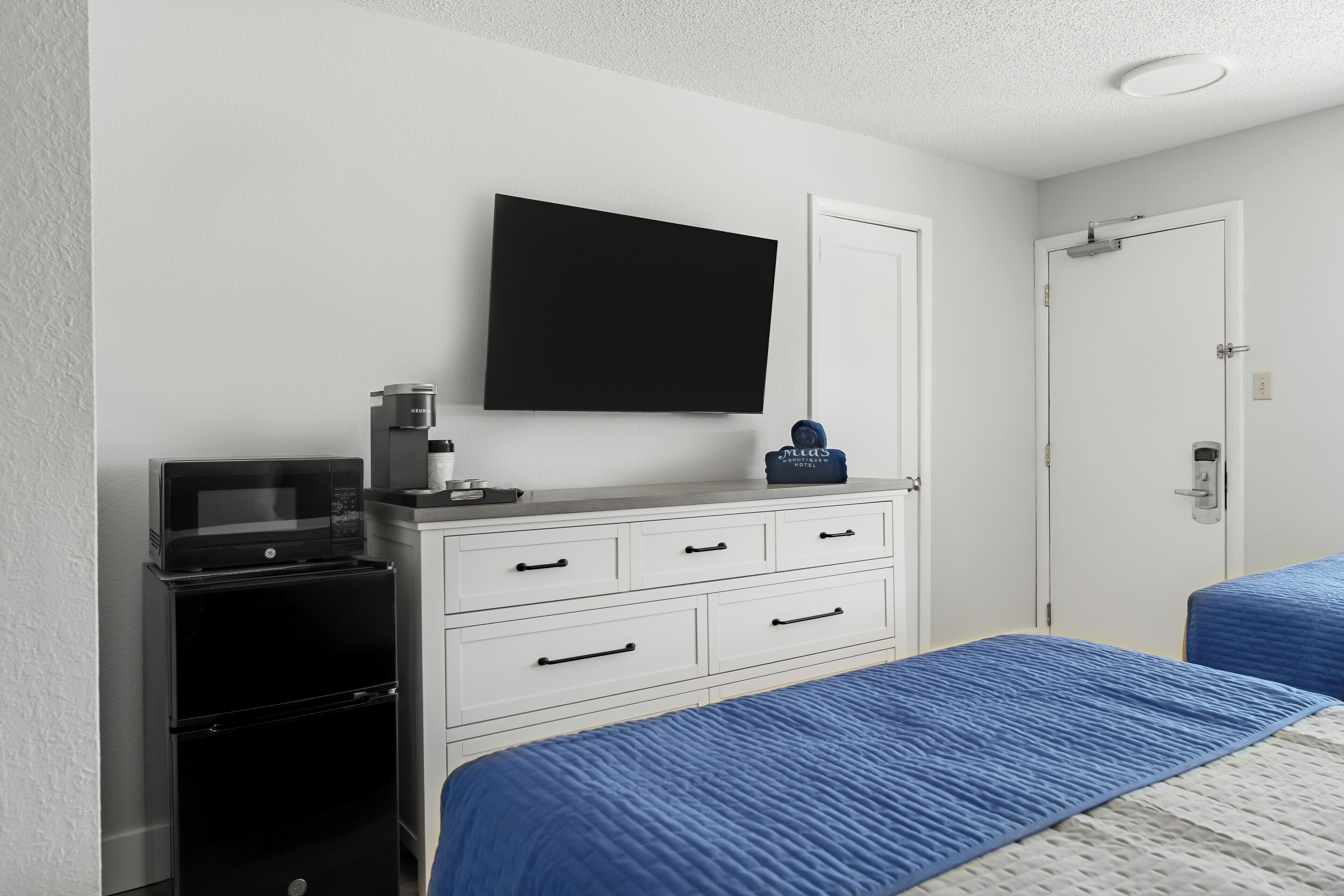 A hotel room with a bed, a desk with a microwave, a flat-screen TV, and a minibar.